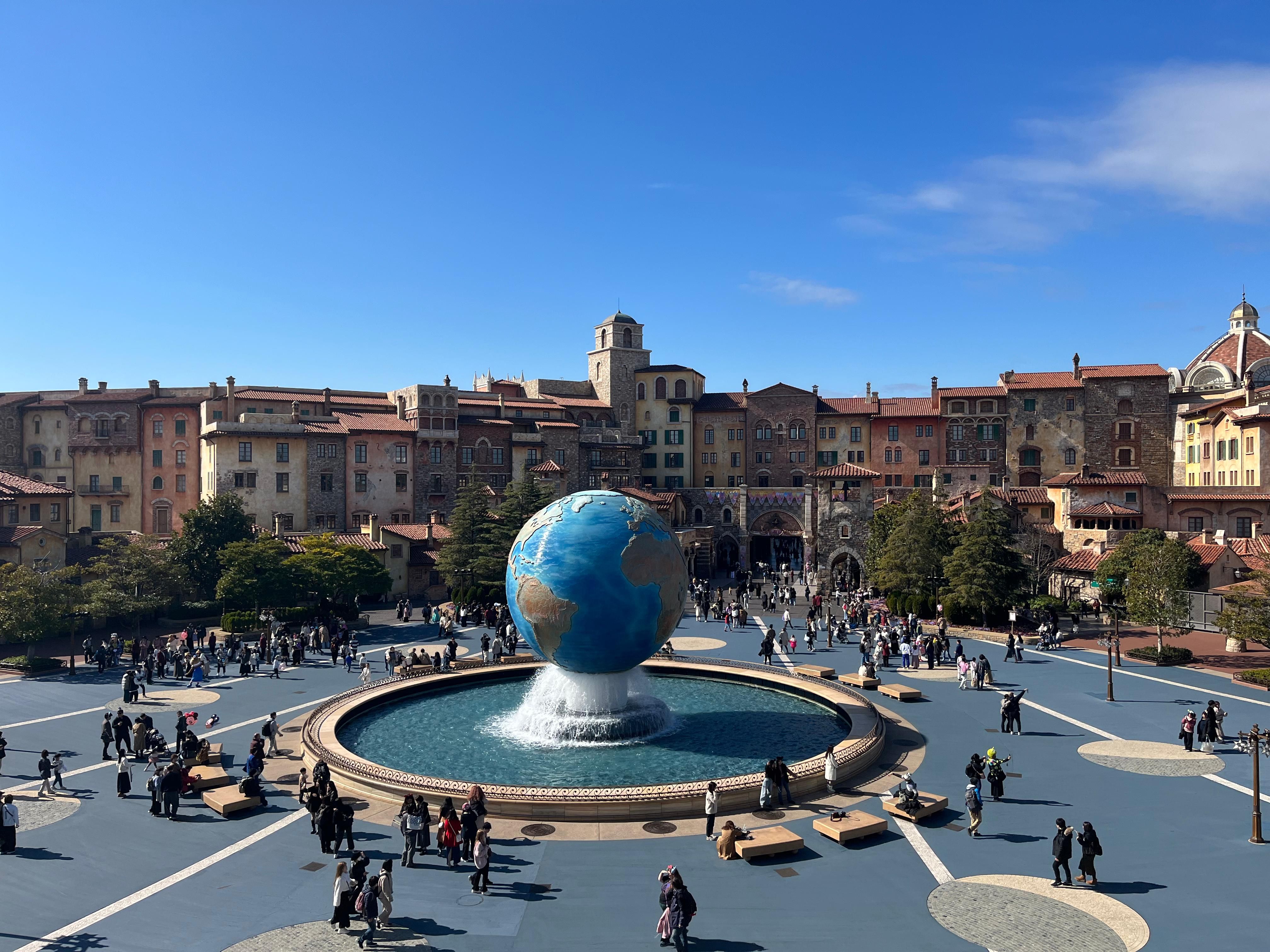 Wide view of a colorful piazza with a central globe fountain, bustling with people under a clear blue sky.