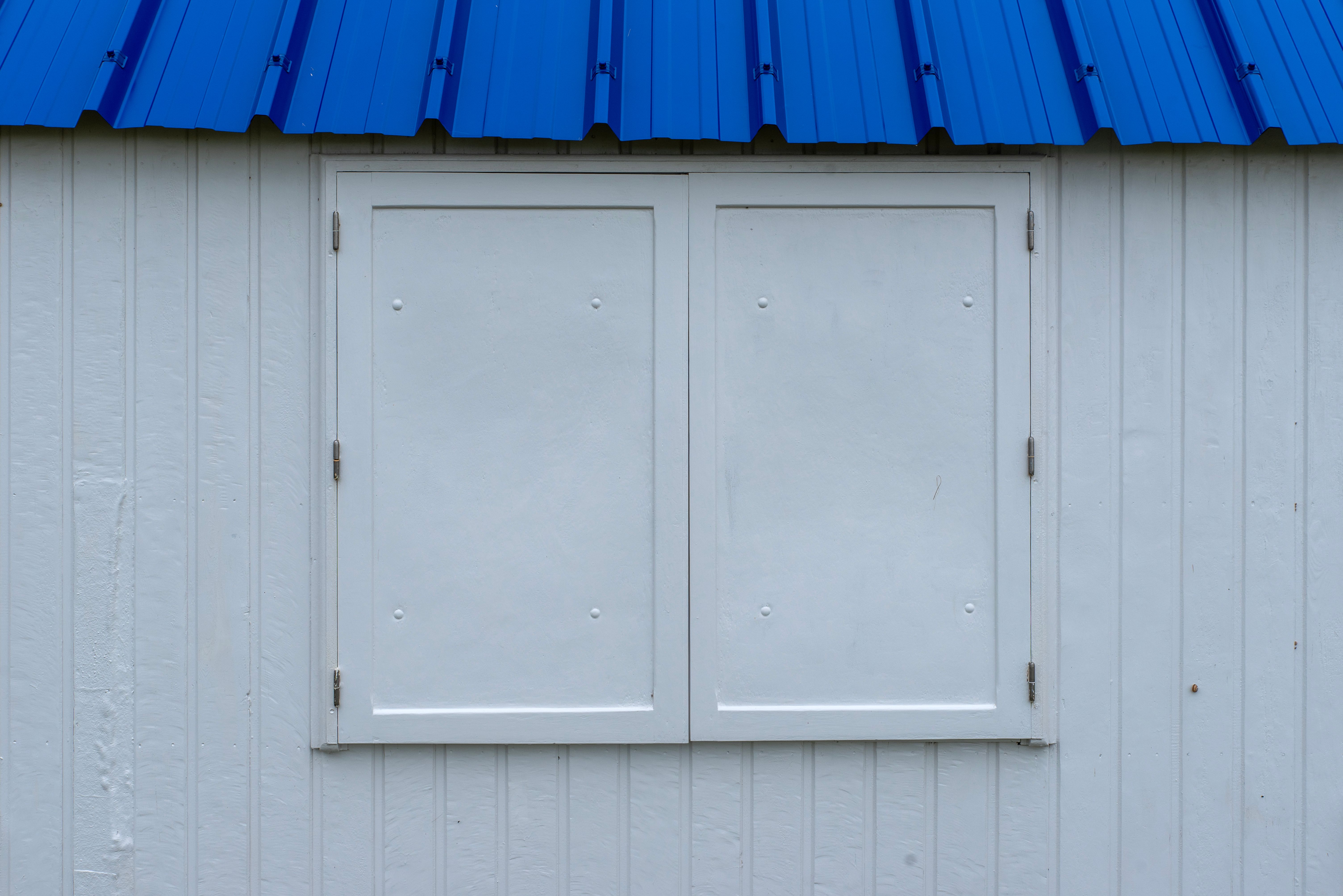 Close-up shot of a white cottage wall with a bright blue roof, featuring boarded-up windows.