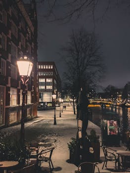 Snow-covered Amsterdam canal at night with illuminated street lamps creating a serene winter scenery.