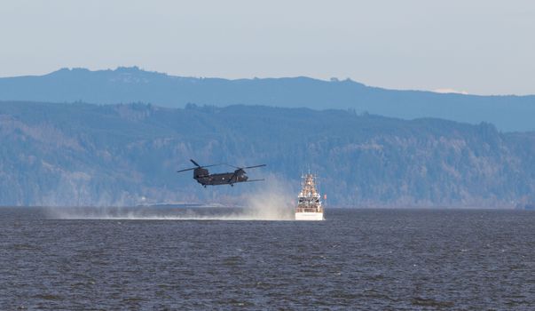 Chinook helicopter flying over water near a ship with forested mountain backdrop.