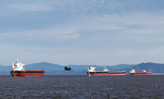 Panoramic view of multiple cargo ships and a helicopter in the sea with mountains in the background.