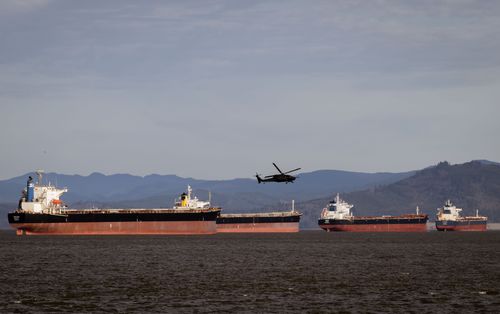 Free A helicopter flies over several cargo ships in a vast ocean with a mountainous backdrop. Stock Photo