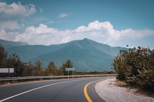 A winding road curves through lush greenery with a mountain backdrop under a bright blue sky.