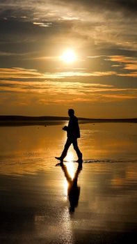 Silhouette of an adult walking on a reflective beach at sunset, creating a dramatic scene.