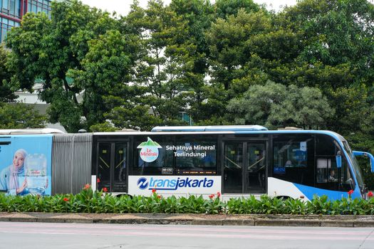 TransJakarta bus passing through a green urban park area with advertisement