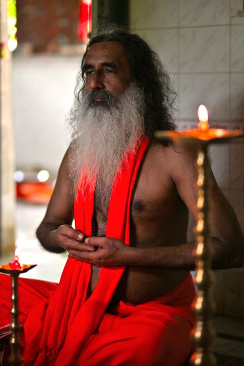Free An elderly guru in meditation, draped in red robes, surrounded by warm candlelight indoors. Stock Photo