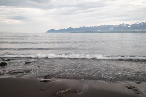 Free Calm seas meet a rocky coast under a cloudy sky with distant snow-capped mountains. Stock Photo