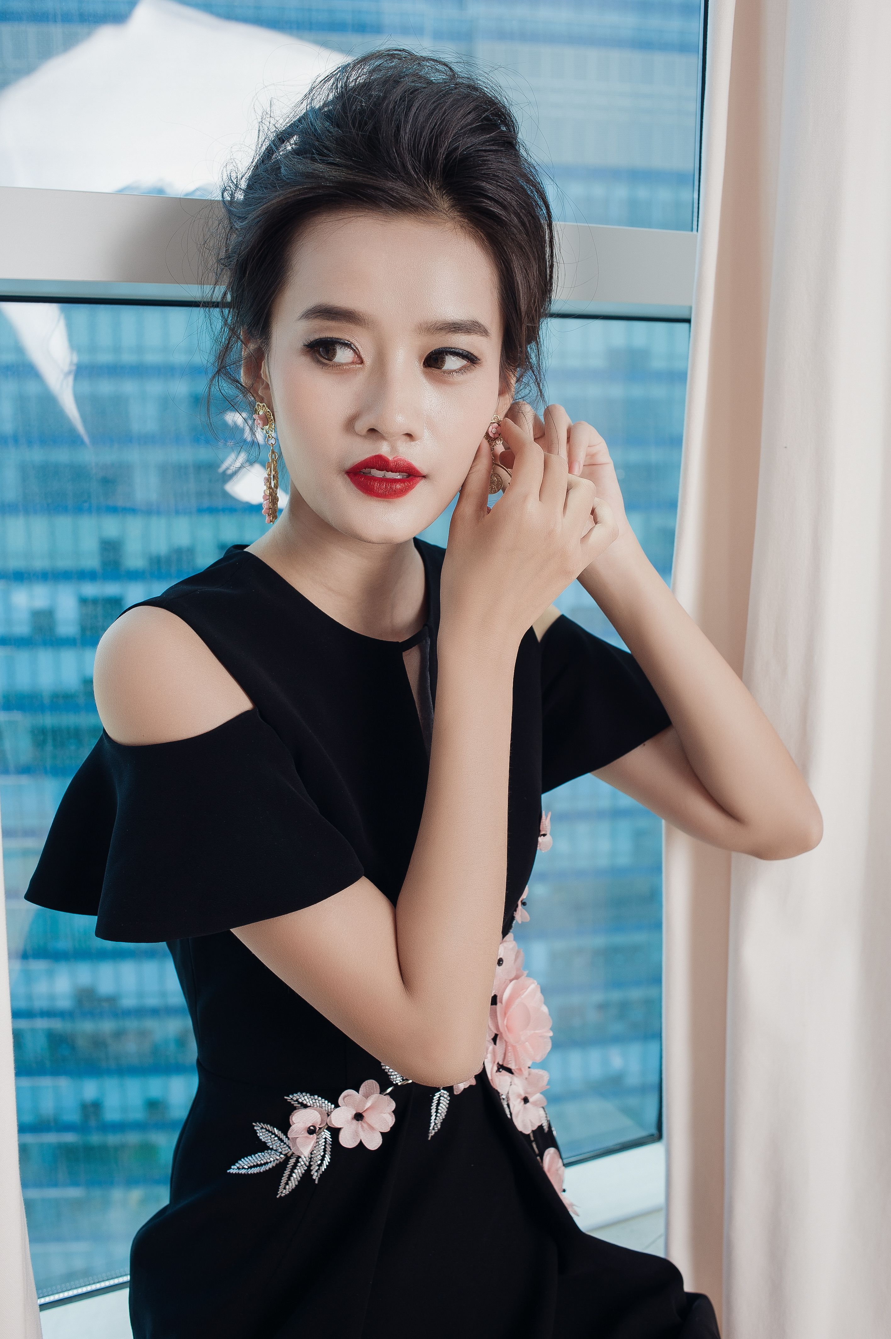 Elegant woman in black dress adjusts earrings near window.