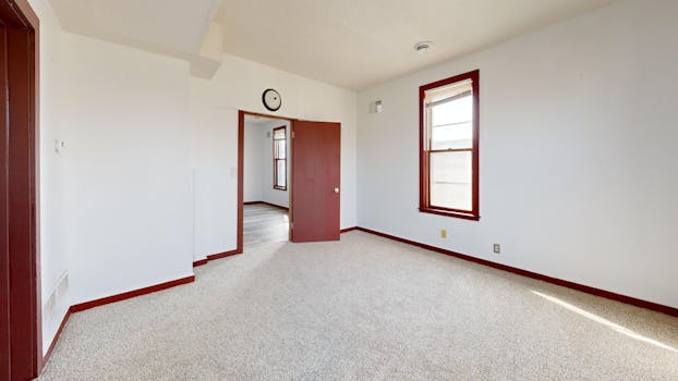 A spacious empty room with red trim, beige carpet, and natural light.