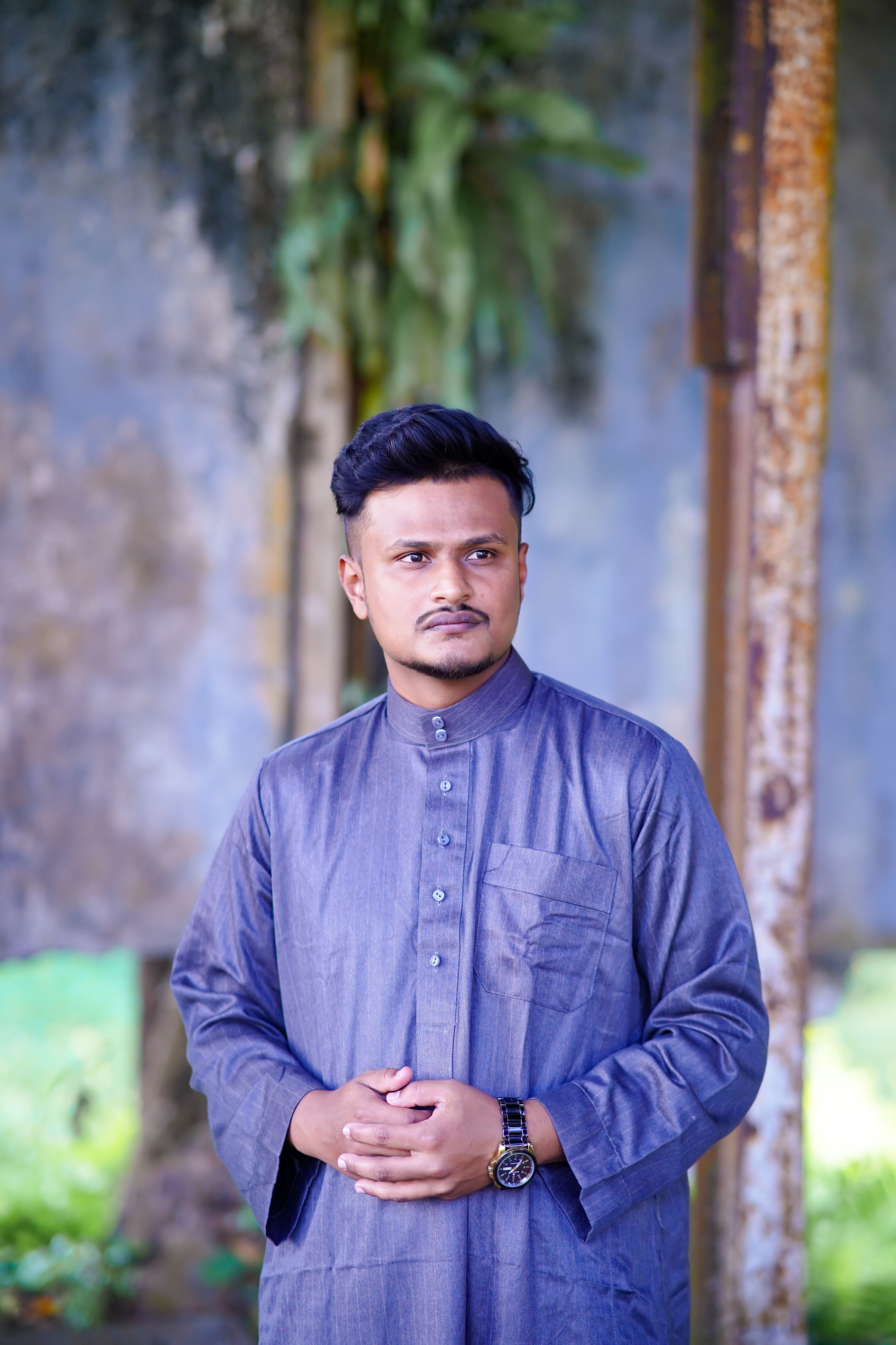 A man in traditional clothing stands outdoors against a rustic backdrop, conveying a contemplative mood.