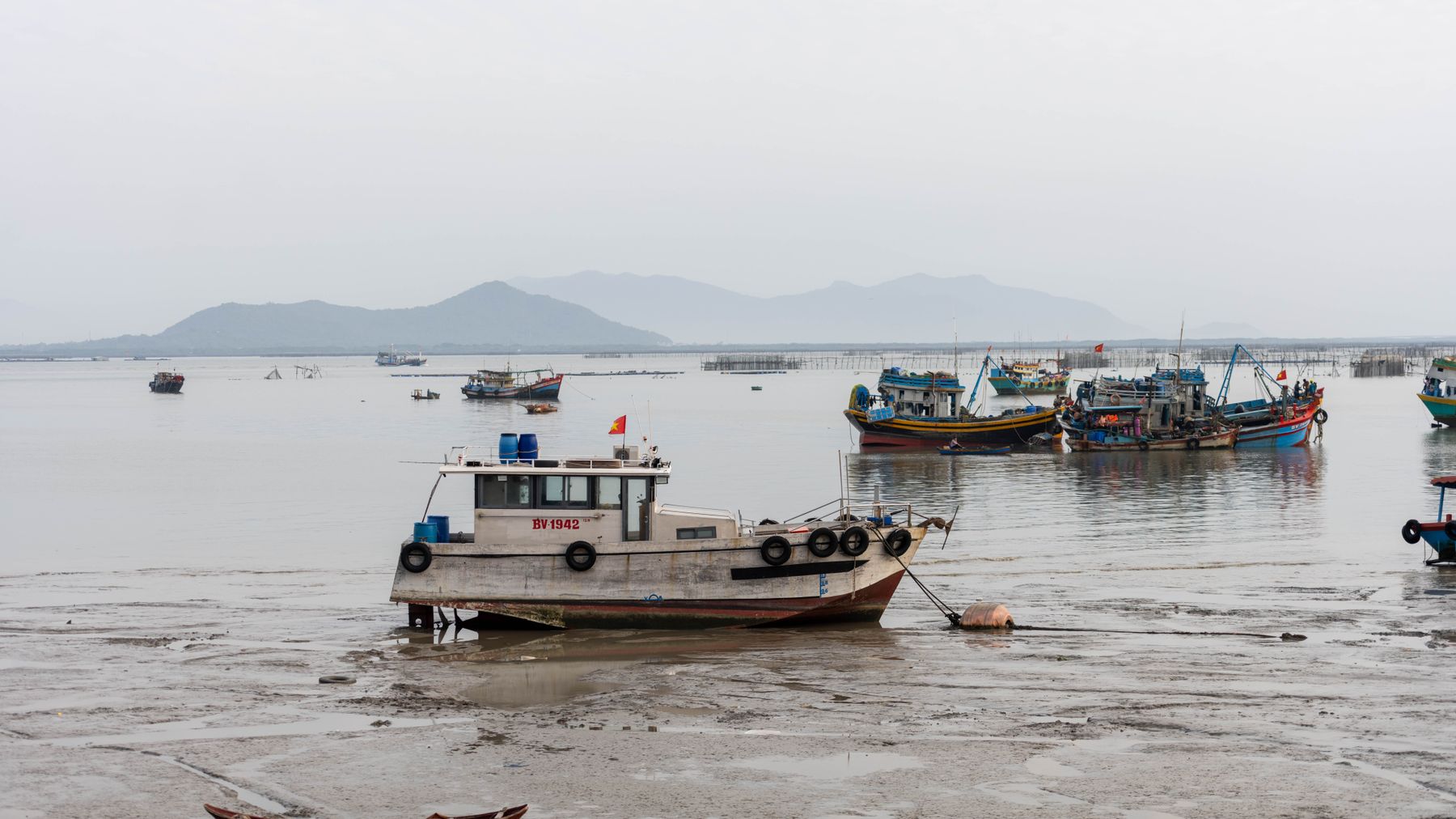 Traditional fishing boats stranded at low tide in Vũng Tàu, Vietnam, with misty mountains in the background.