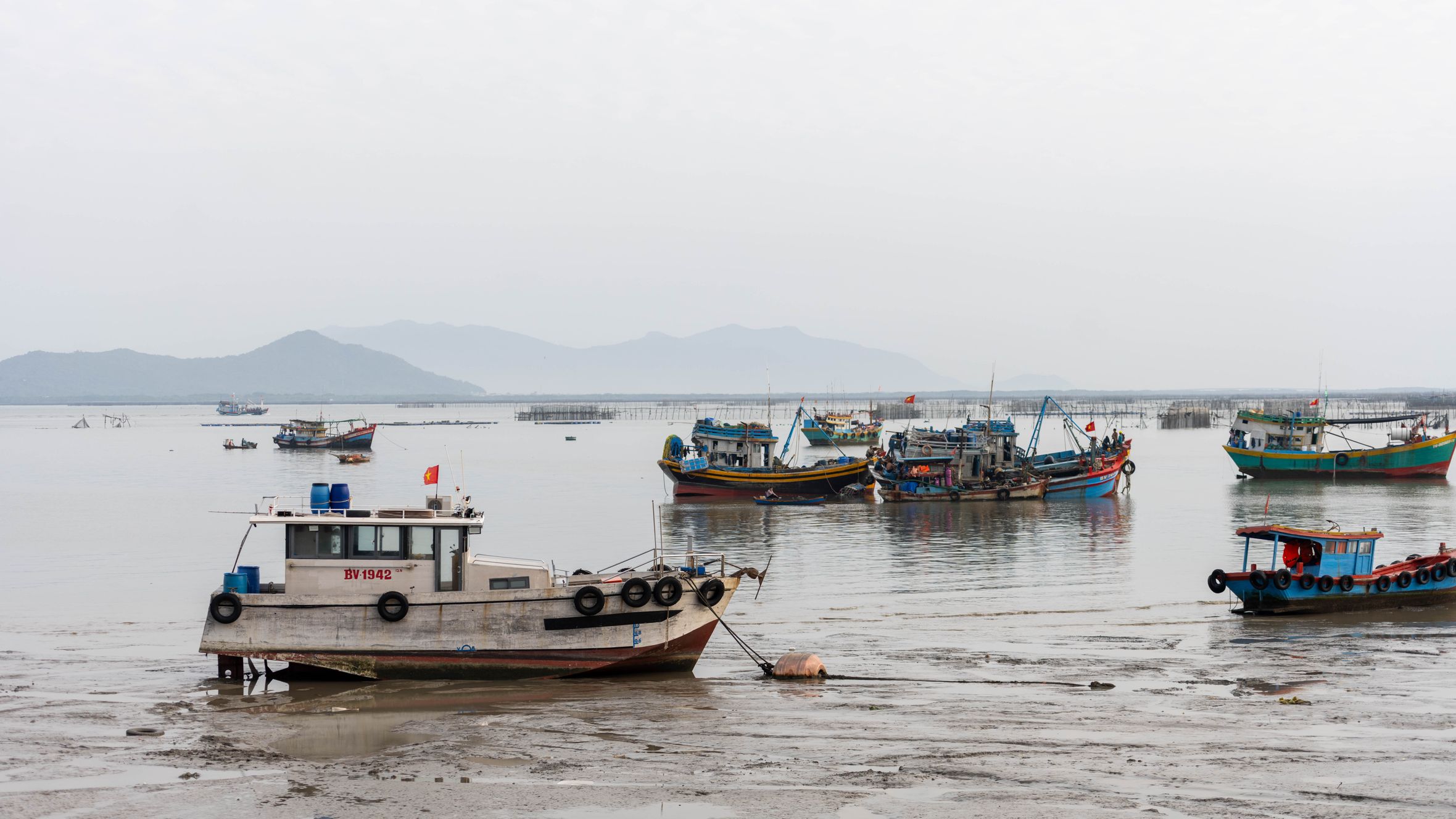 Colorful fishing boats docked at low tide in Vũng Tàu, Vietnam with a mountainous backdrop.