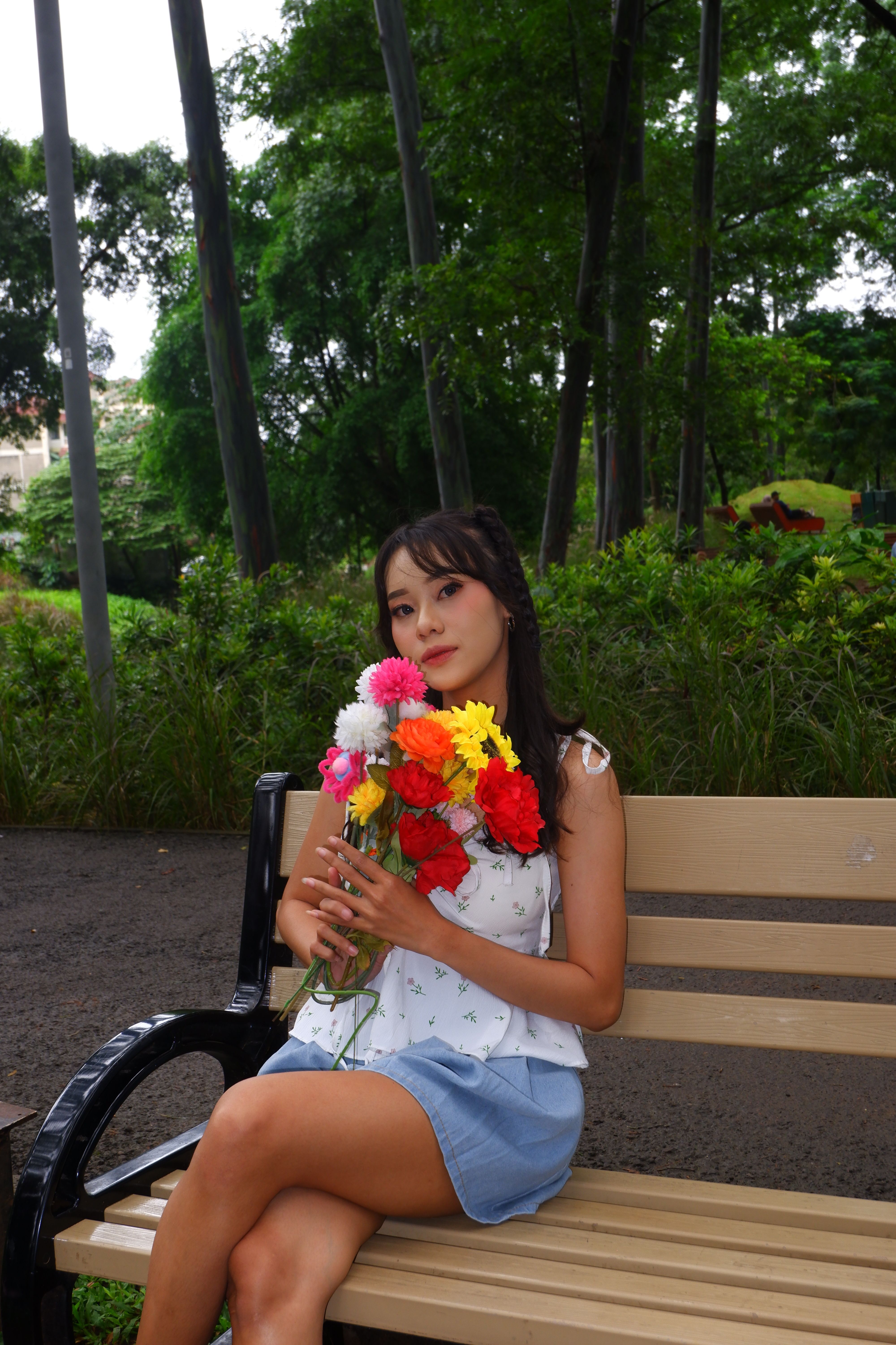 A young woman sits on a park bench in Jakarta, holding a vibrant bouquet of flowers.