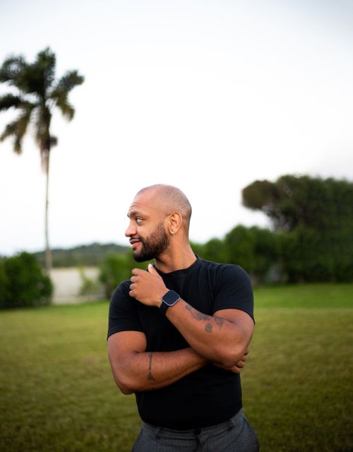 Free Bald man with a beard standing outdoors, casually dressed in a black shirt, surrounded by greenery at dusk. Stock Photo