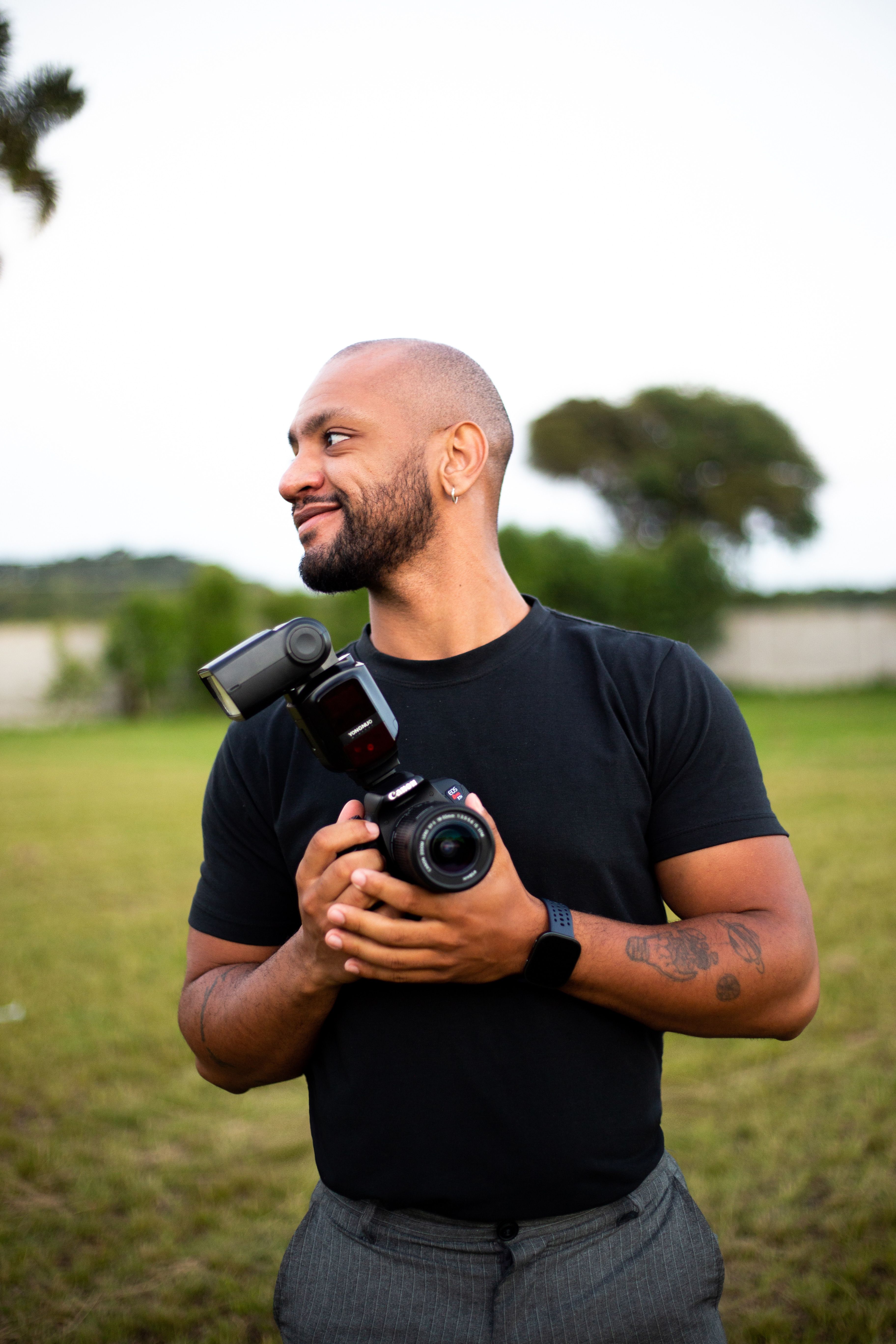 A smiling adult male photographer holding a camera in an outdoor setting with greenery.