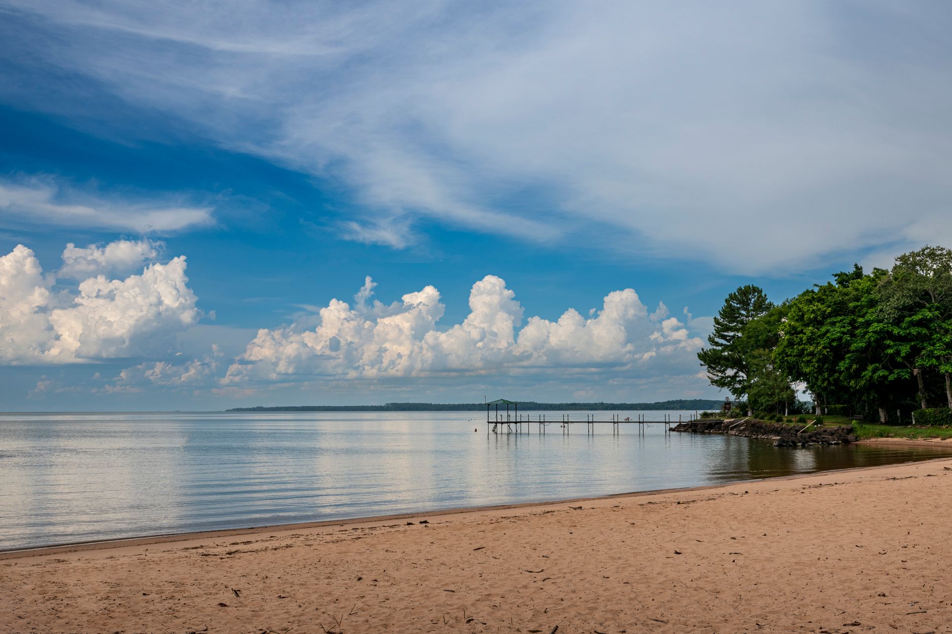 Tranquil beach scene with a pier, calm waters, and cumulus clouds under a blue sky.