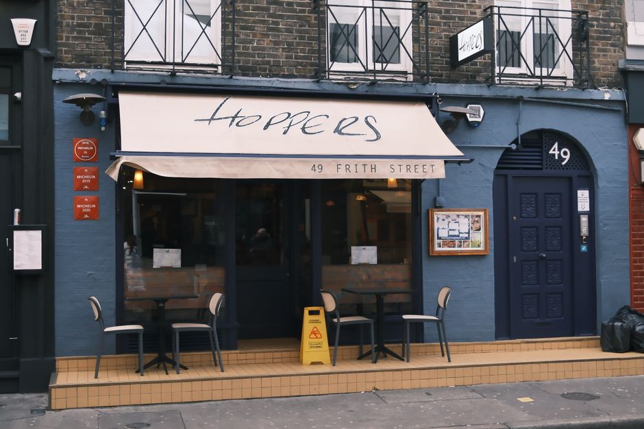 Front view of Hoppers, a cozy restaurant on Frith Street, London, featuring outdoor seating and Michelin signs.