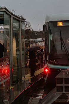 Passengers waiting with umbrellas at a rainy Istanbul tram stop during twilight.