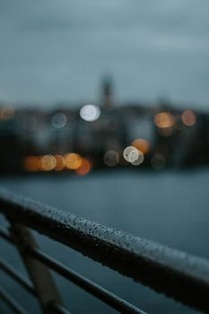 Blurred city lights of İstanbul with wet railing foreground, evoking a moody atmosphere.