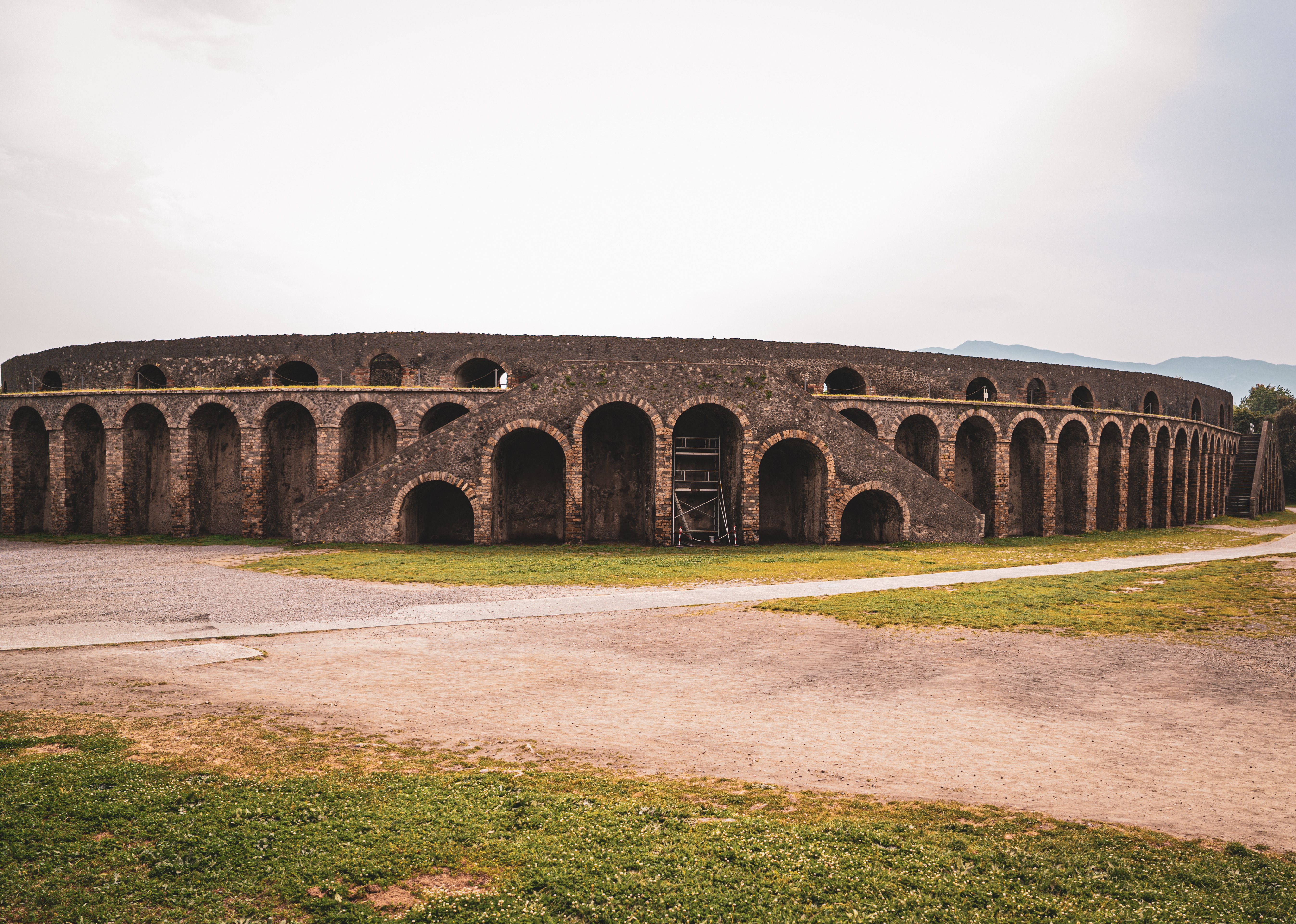 Ancient brick amphitheater arches in Pompeii, Italy, with grassy ruins, distant hills, no people, and soft overcast light