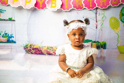 Cute baby girl in white dress celebrates birthday indoors with colorful decorations.