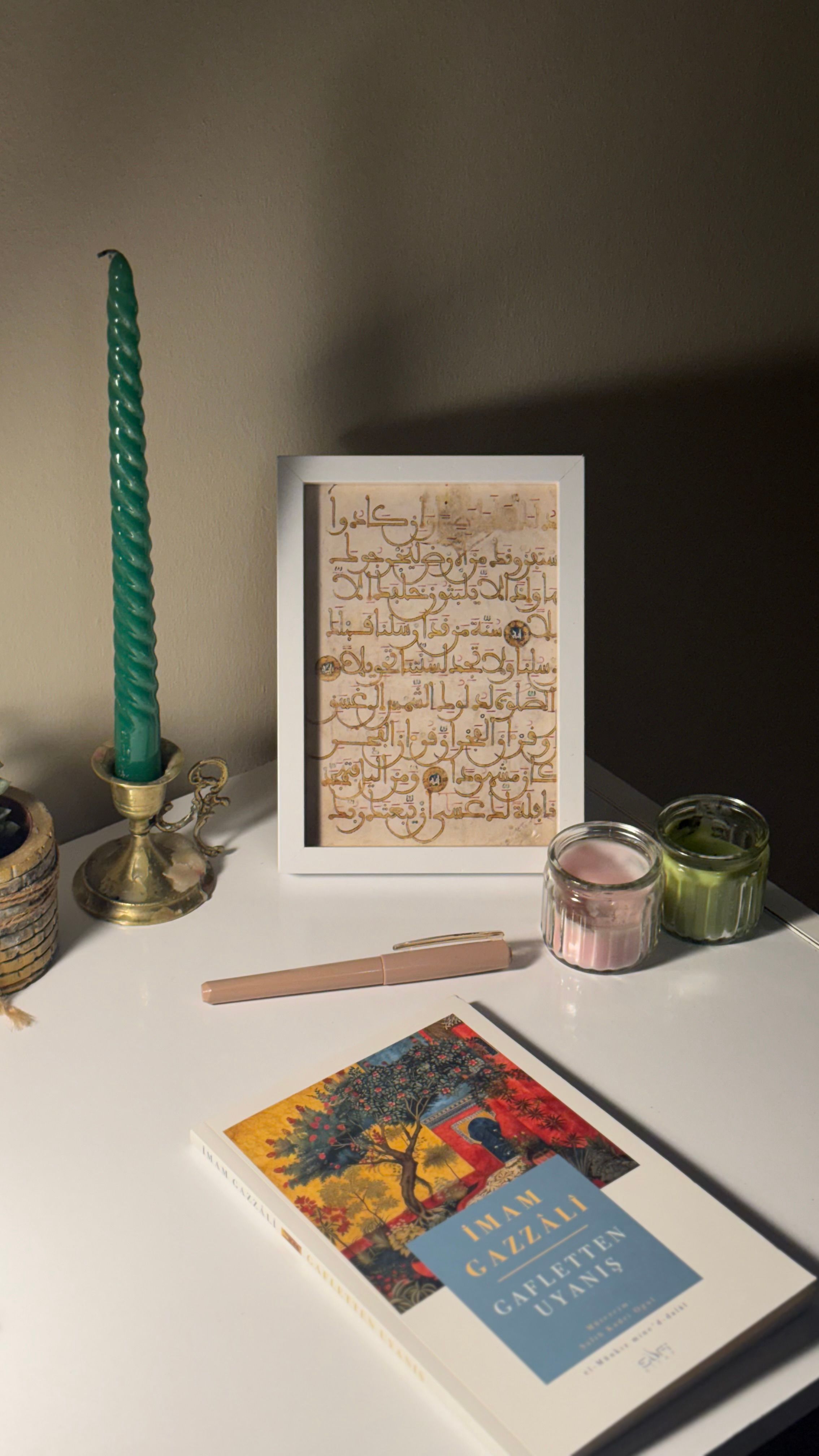 A serene tabletop featuring a book, green candle, framed Arabic calligraphy, and decorative jars.