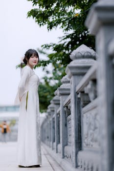 A woman in a white traditional dress posing elegantly outdoors near a stone railing.