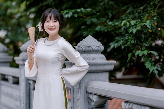 Donna sorridente in ao dai bianco che tiene un ventaglio, in piedi all'aperto.