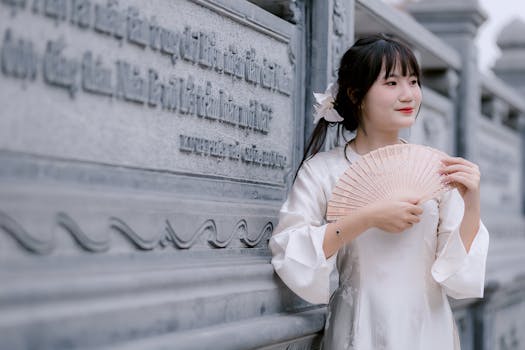 A young woman in traditional attire holding a fan near an ornately carved stone wall.