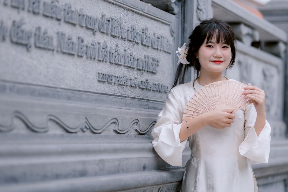 Young woman in traditional Asian dress holding a fan, standing by ornate stone wall.