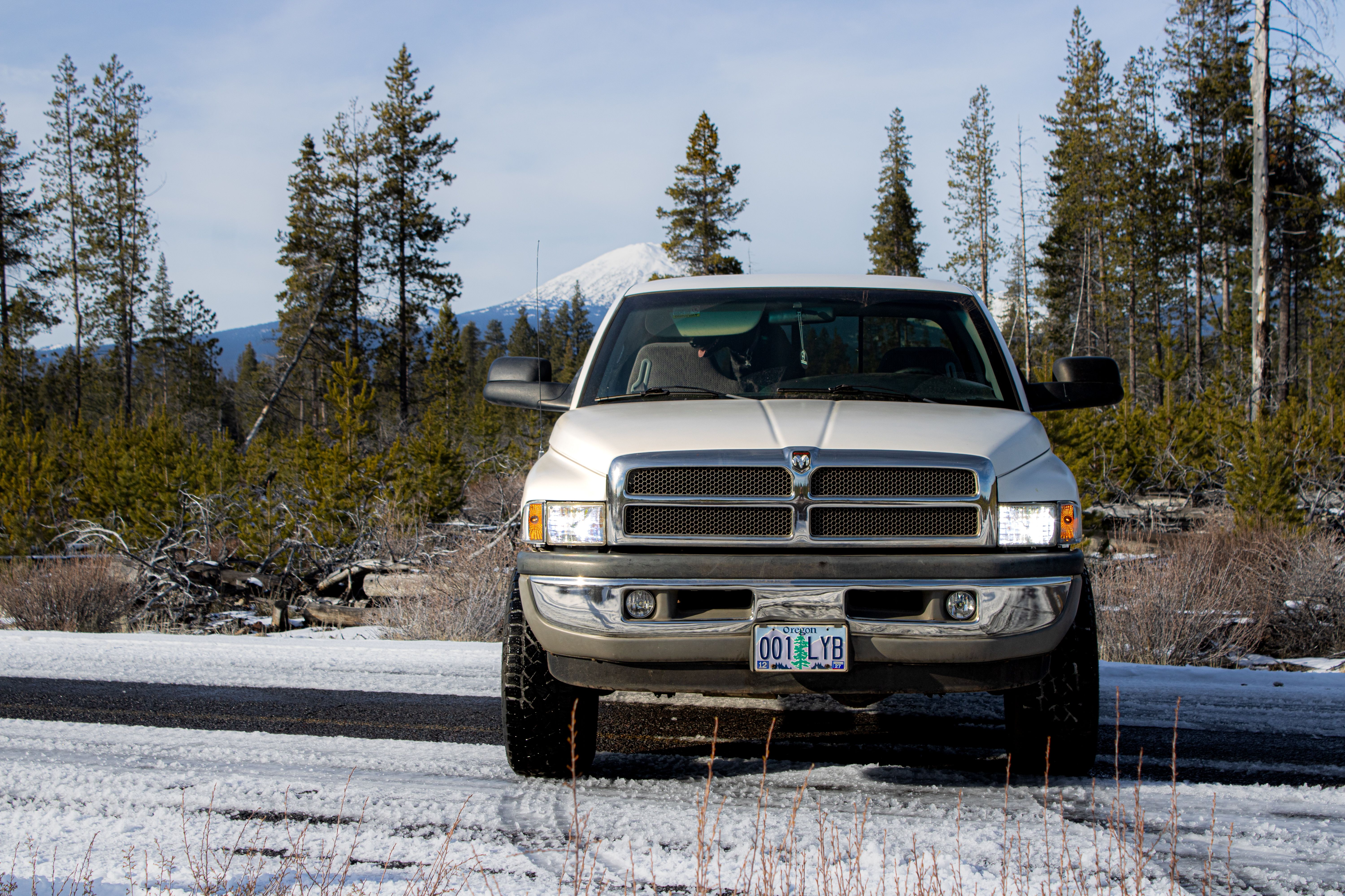 A truck parked on a snowy road with a forest and mountain backdrop under a clear blue sky.