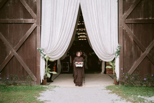 A woman in a brown dress holding flowers stands at the entrance of a rustic wedding venue with open curtains.