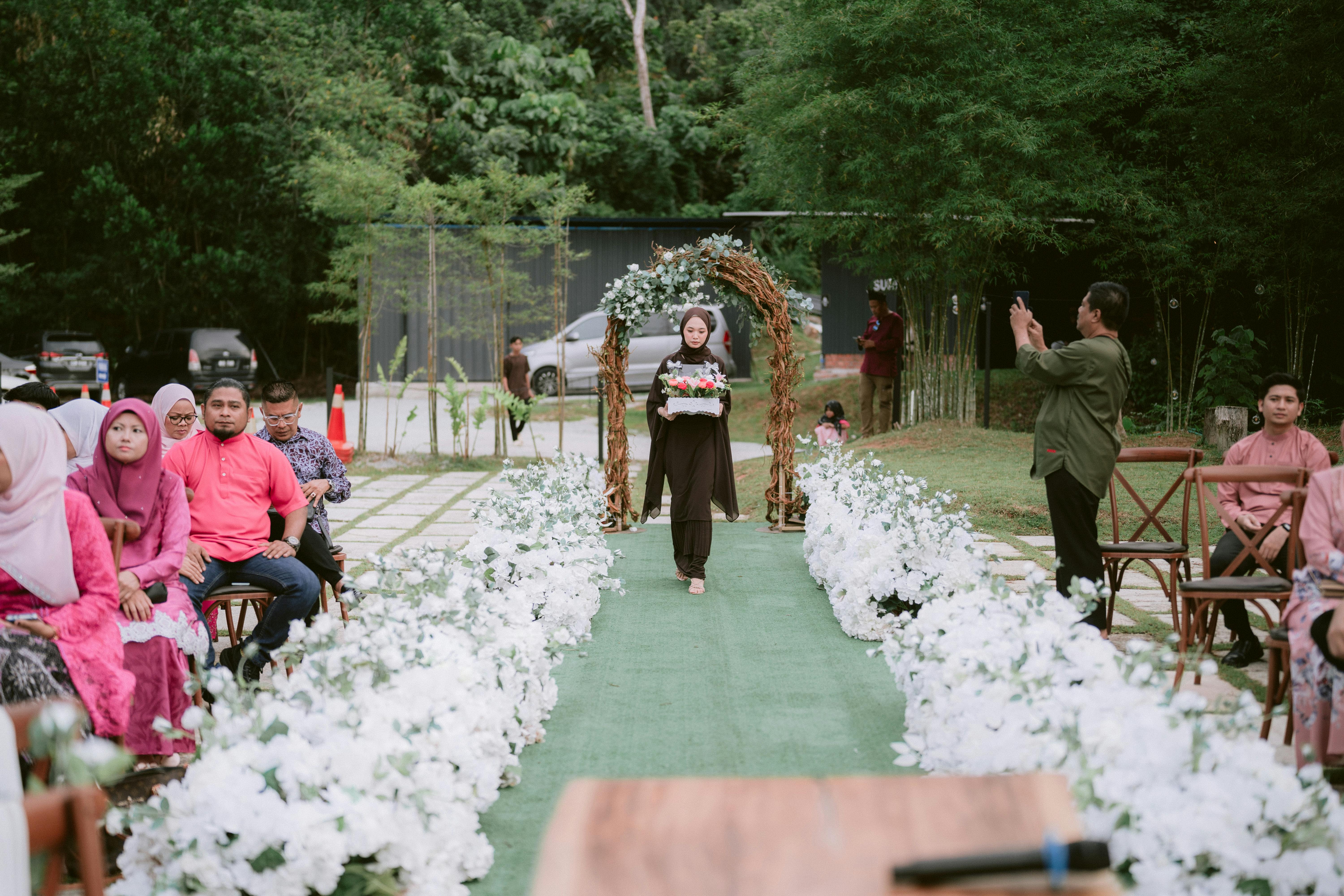 Outdoor wedding ceremony with guests and floral decorations. A woman walks down the aisle.