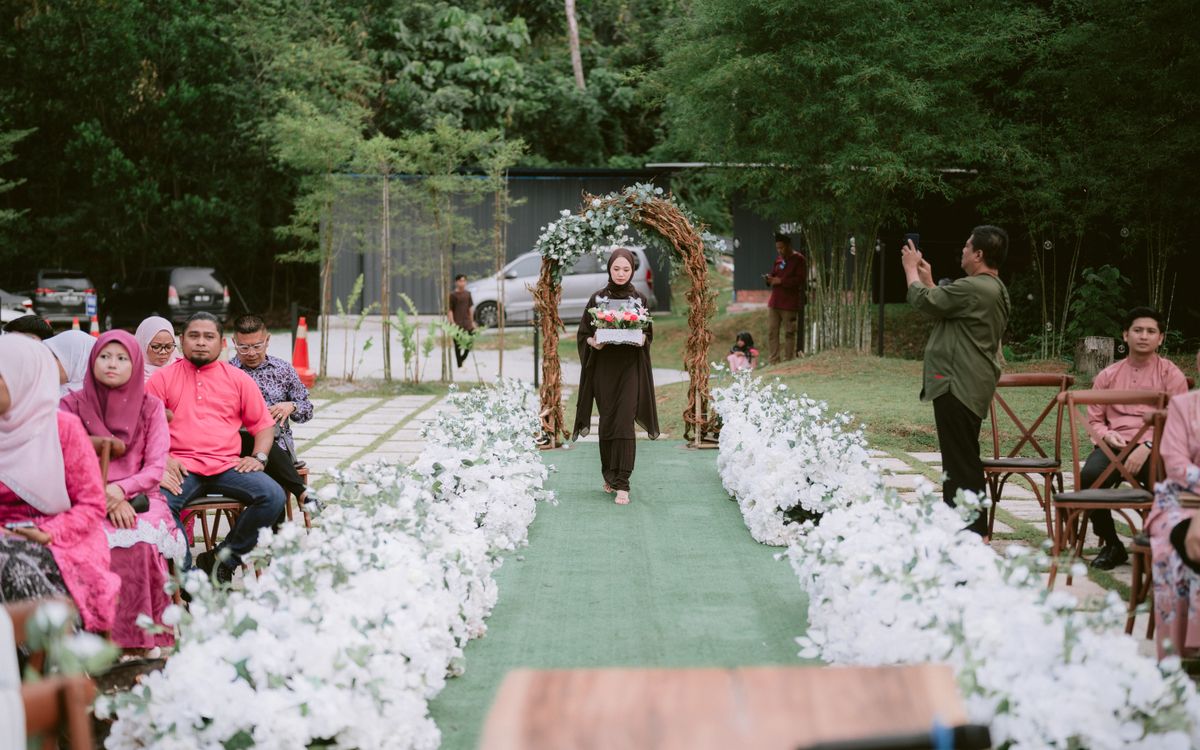 Outdoor wedding ceremony with guests and floral decorations. A woman walks down the aisle.