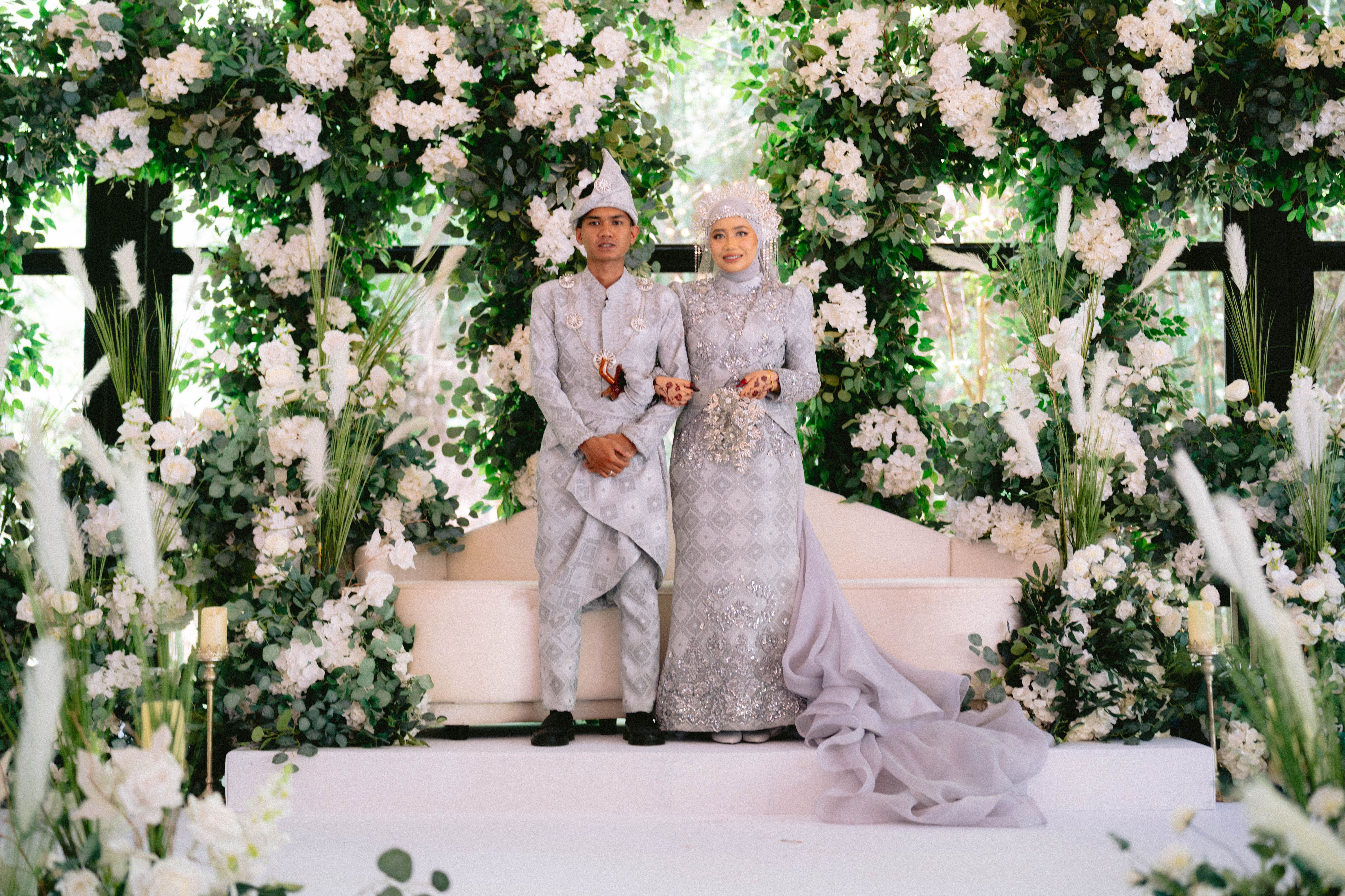 A couple in traditional attire poses amid lush floral arrangements at their wedding ceremony.