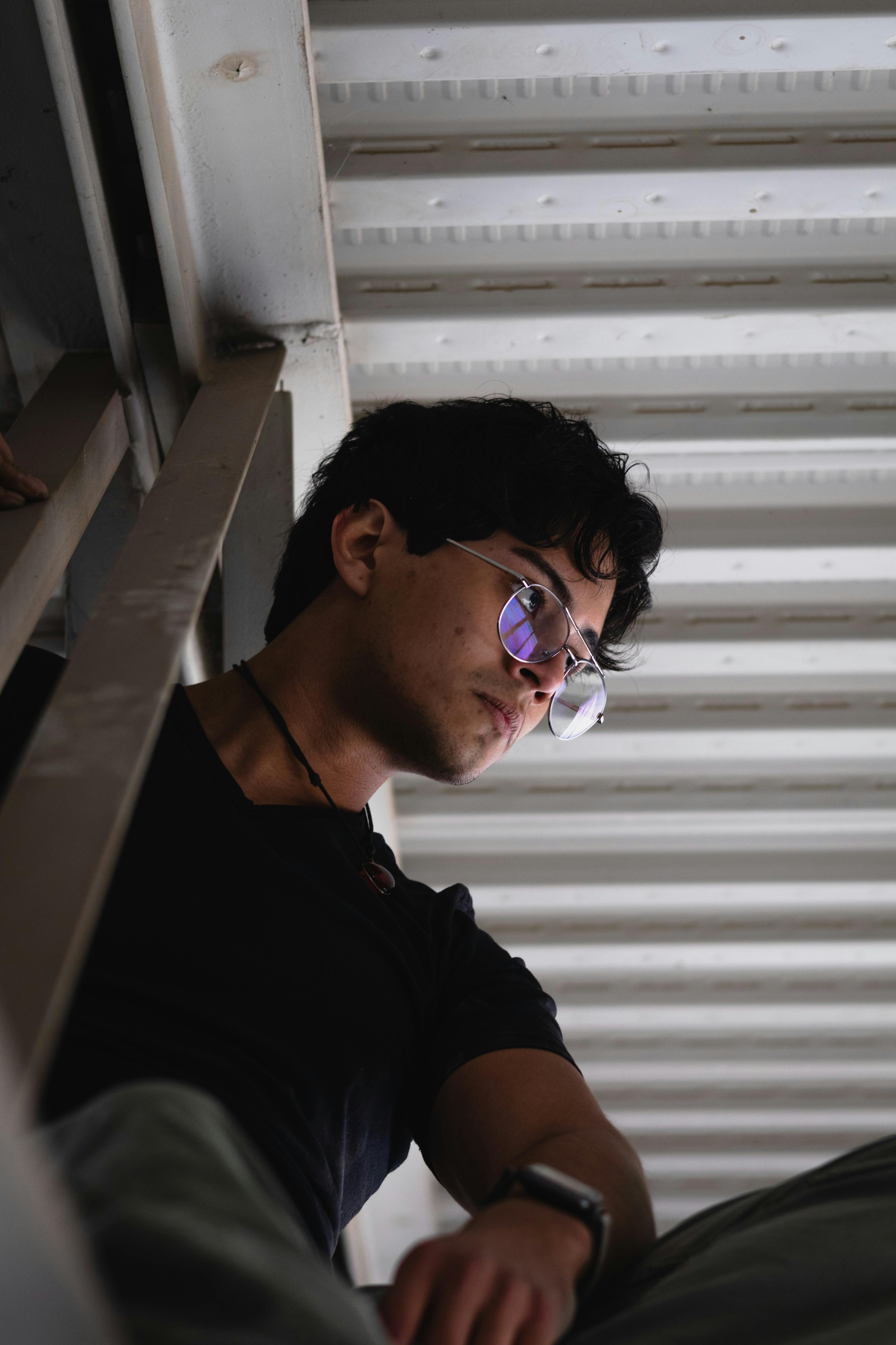 grátis Jovem de óculos, absorto em pensamentos, sob vigas industriais expostas, evocando a vida urbana. Foto profissional