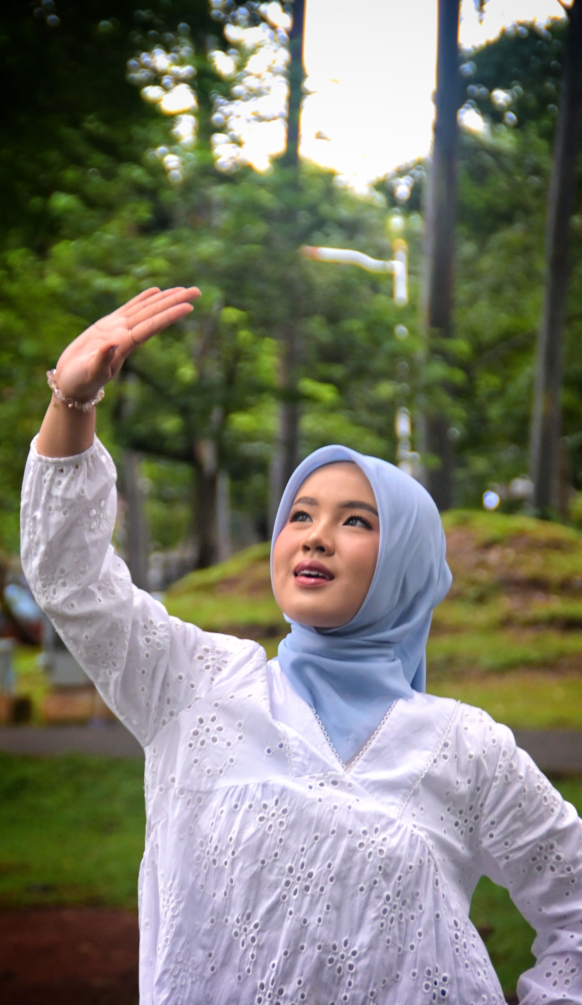 A young woman in a blue hijab and white dress enjoys the outdoors with trees in the background.