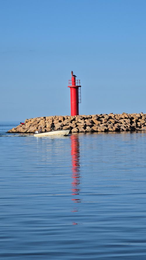 Free Vibrant red lighthouse stands on rocky jetty, reflecting in calm sea under clear blue sky. Stock Photo