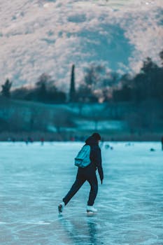 A young adult wearing a backpack ice skating on a frozen lake during winter, surrounded by snowy mountains.