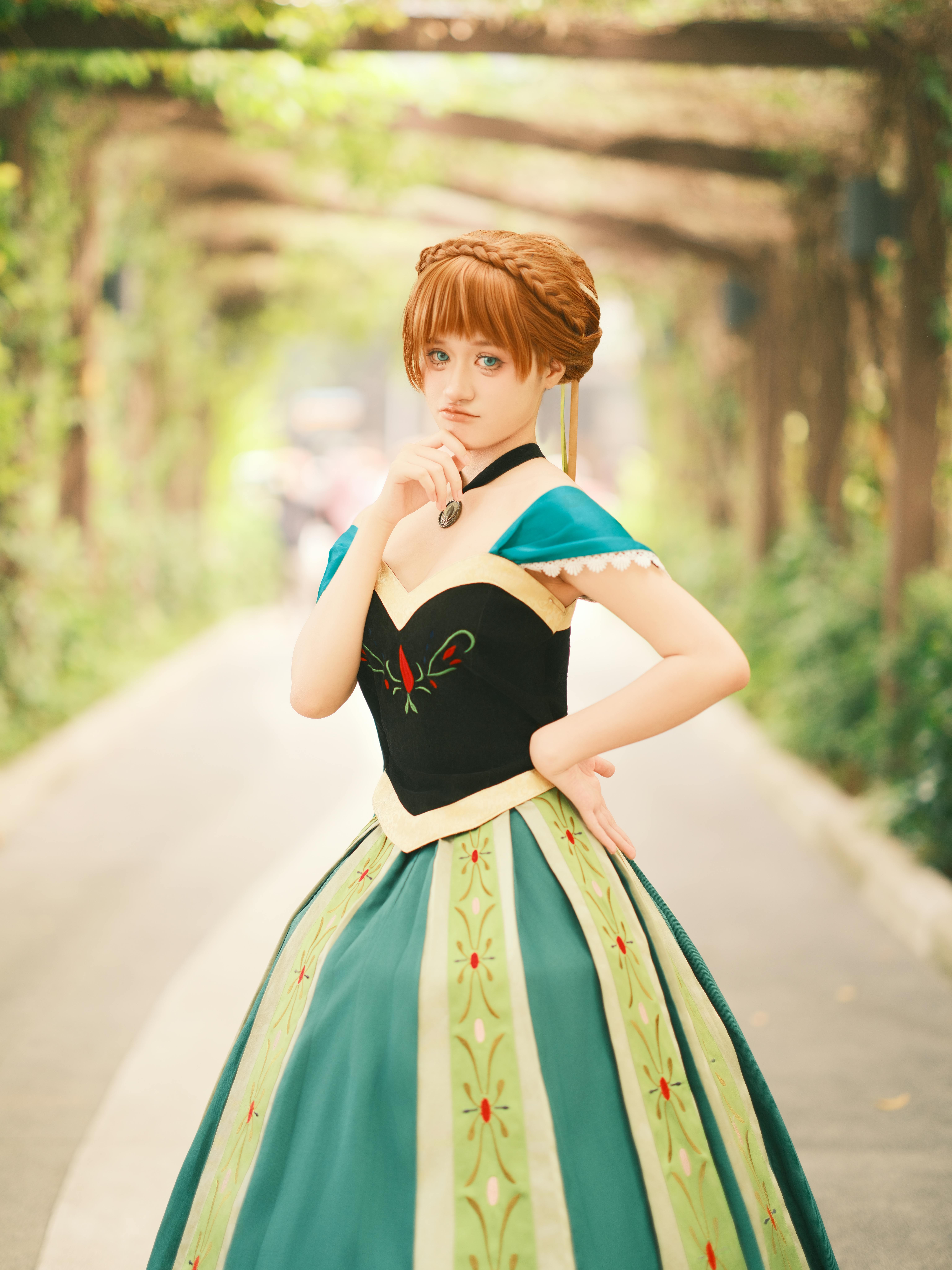 A young woman in princess cosplay poses thoughtfully in a lush, leafy garden pathway.