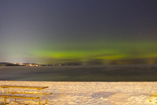 Aurora Borealis illuminates the night sky over Jönköping's snowy beach with a scenic view of the city lights.