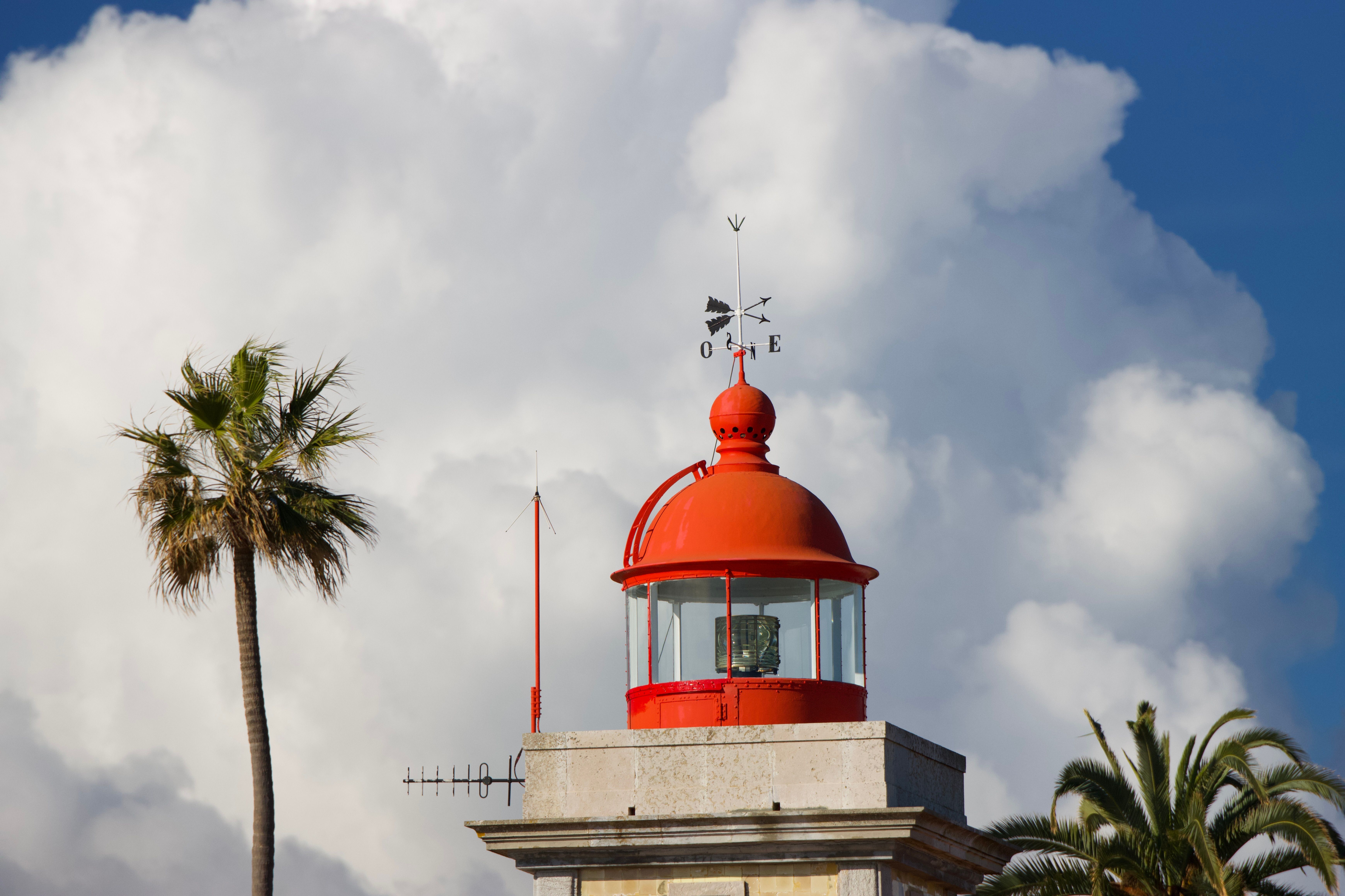 A striking red lighthouse in Lagos, Portugal stands against dramatic clouds and palm trees. - Lagos