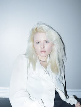 Elegant portrait of a young woman with blond hair in a white outfit against a minimal background.