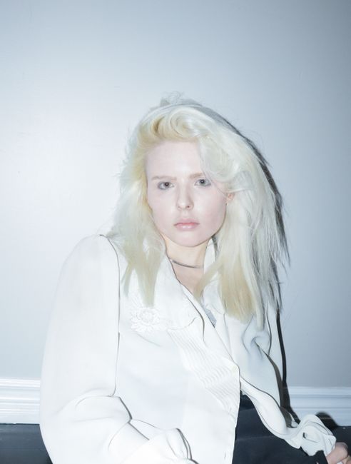 Elegant portrait of a young woman with blond hair in a white outfit against a minimal background.