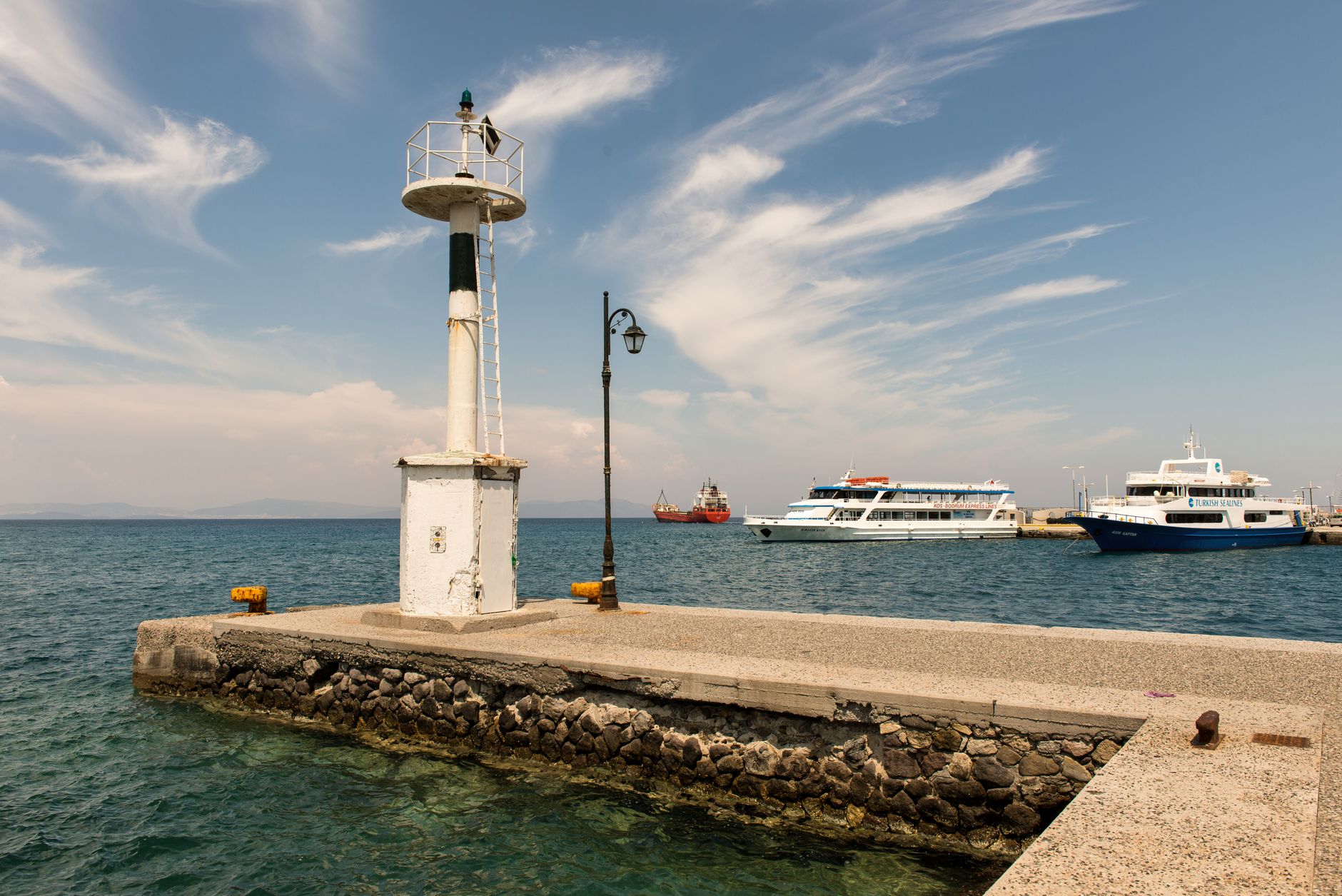 A picturesque view of a lighthouse and boats in a tranquil Greek harbour, under a clear blue sky.