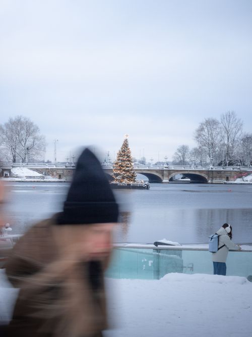 Free A Christmas tree lights up the snowy Alster River in Hamburg with blurred people passing by. Stock Photo