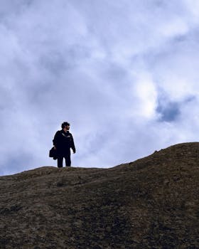 Solitary figure atop a rocky hill under a cloudy sky, symbolizing adventure.