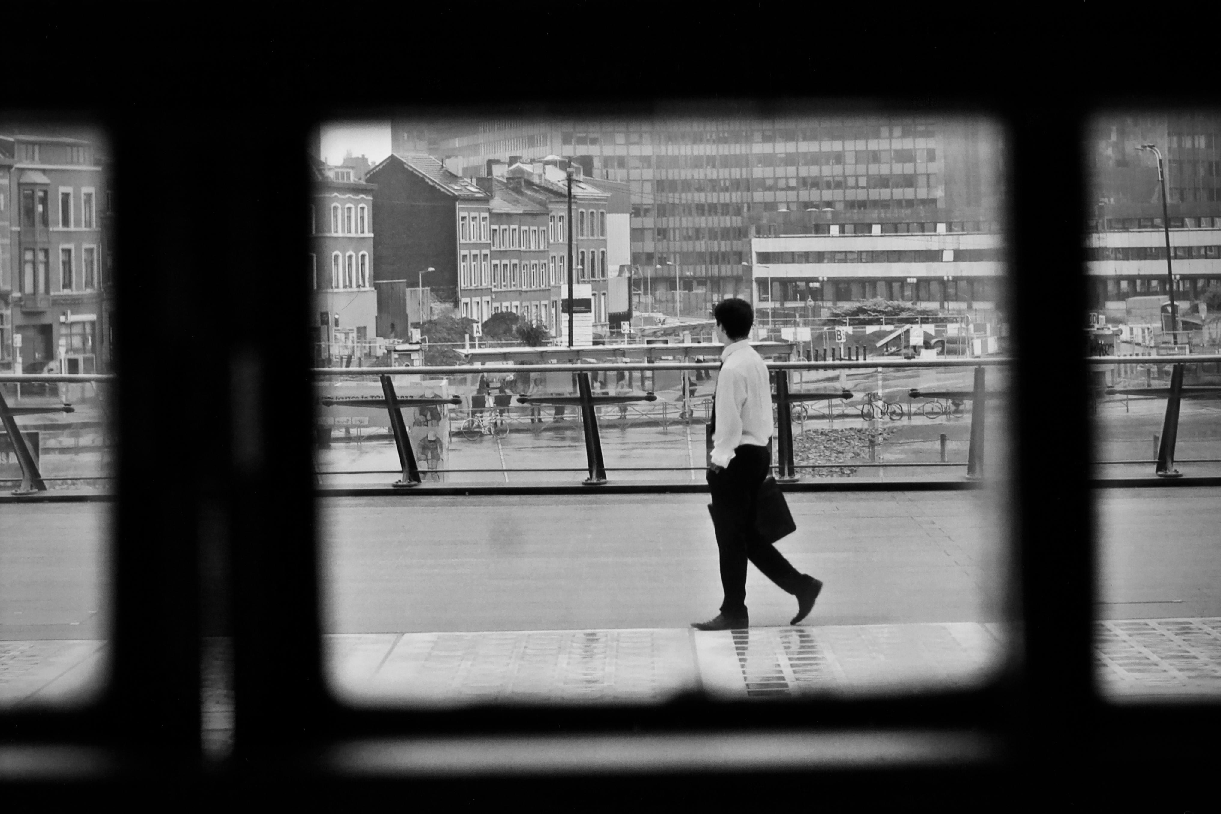 Kostenlos Schwarzweißaufnahme eines Mannes, der auf einem erhöhten Gehweg entlanggeht; im Hintergrund sind Stadtgebäude zu sehen. Stock-Foto