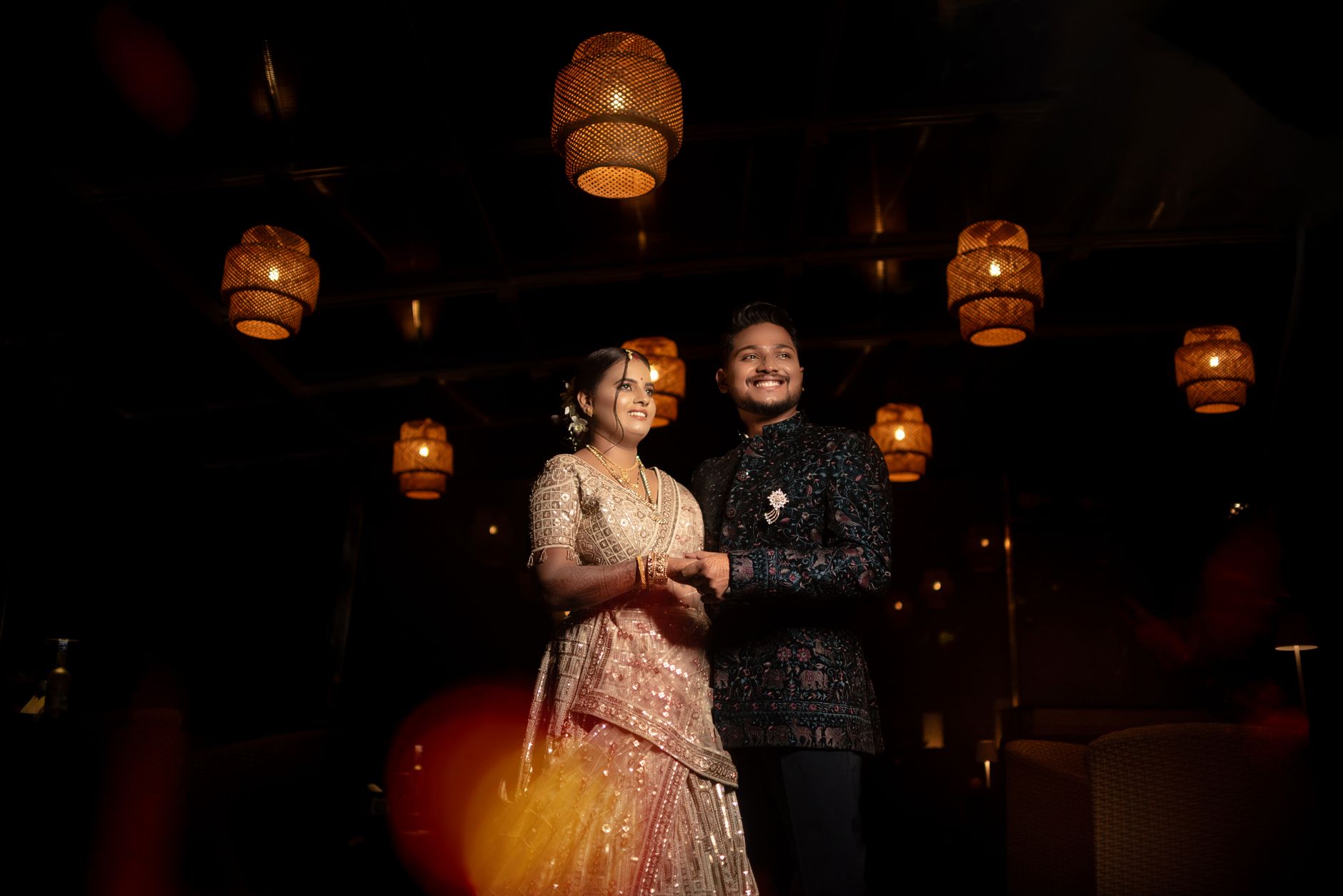 A couple celebrates their Indian wedding, dressed in traditional attire, under warm glowing lights.