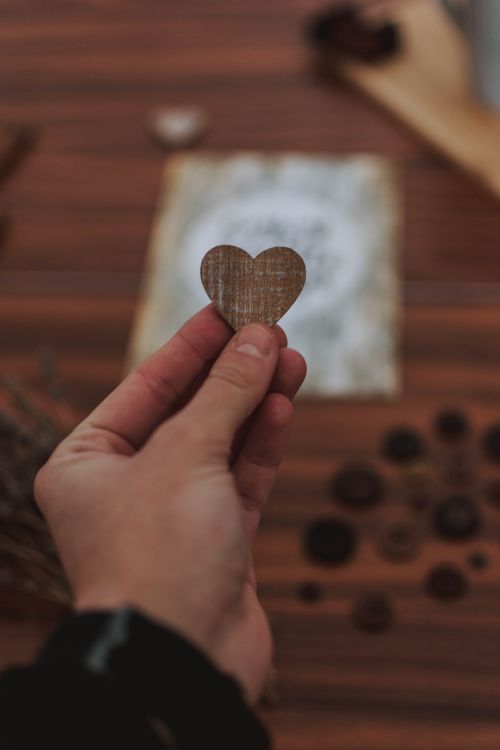 Free Close-up of a hand holding a wooden heart-shaped ornament with a rustic table setting. Stock Photo